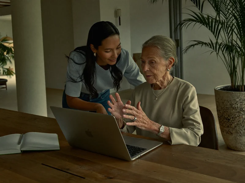 Donna anziana al computer con quaderno, affiancata da un tutor attento